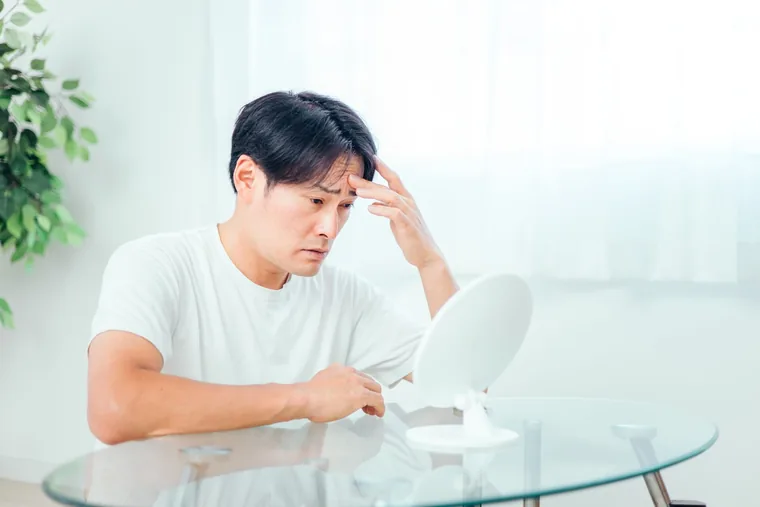 A man in his forties looking closely at his face in a hand mirror, touching his forehead